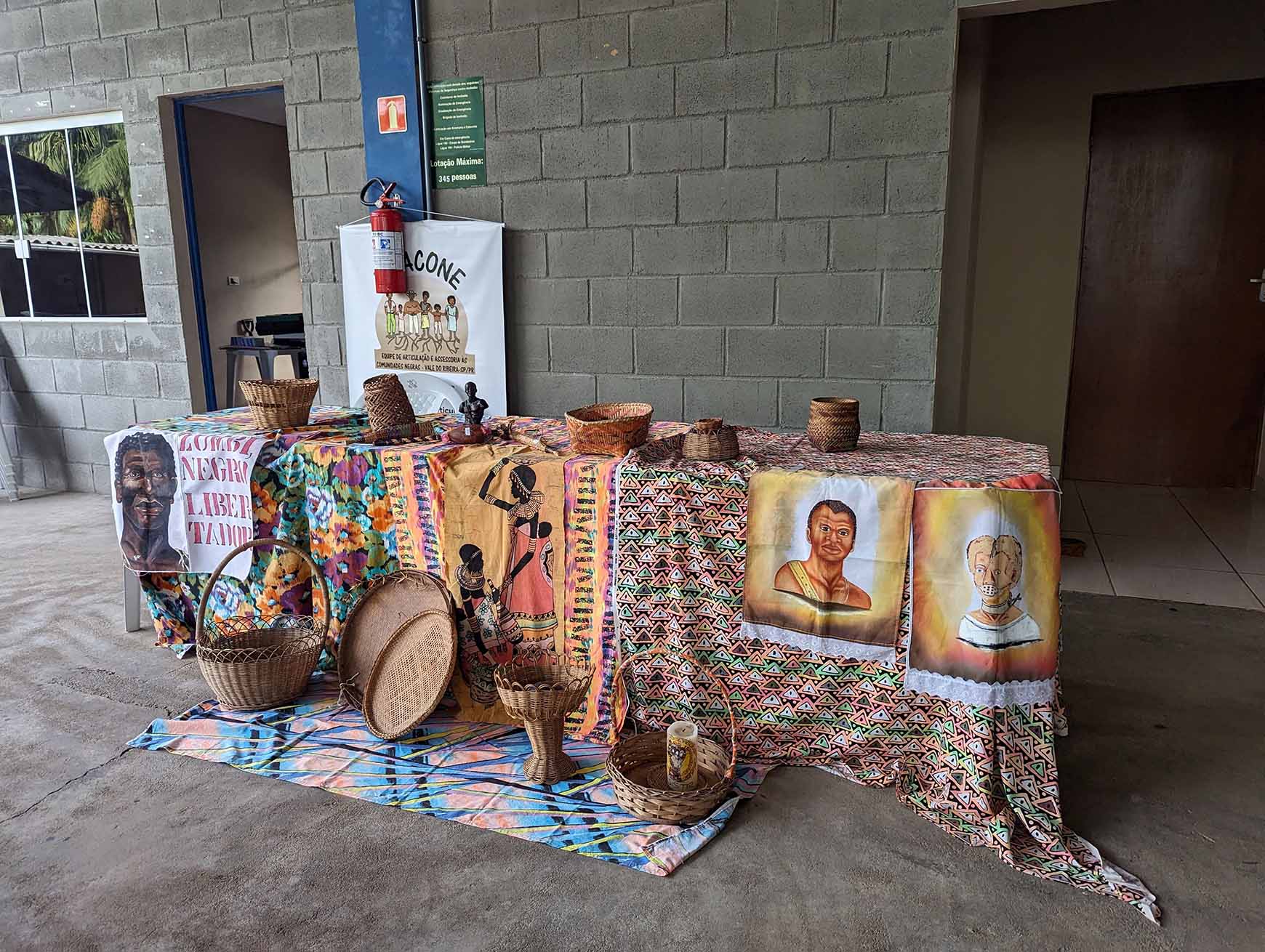 Image of a display table at a quilombo communities meetup in Eldorado, Vale do Ribeira, São Paulo, Brazil. Table draped with textile, featuring basketry and folk images. 