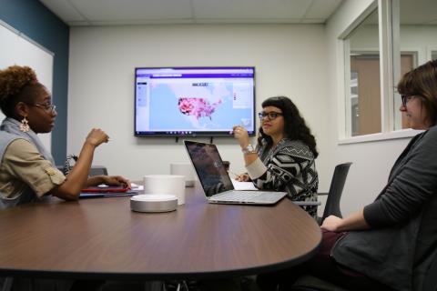 three people discussing a project at a table with computer monitor on the wall