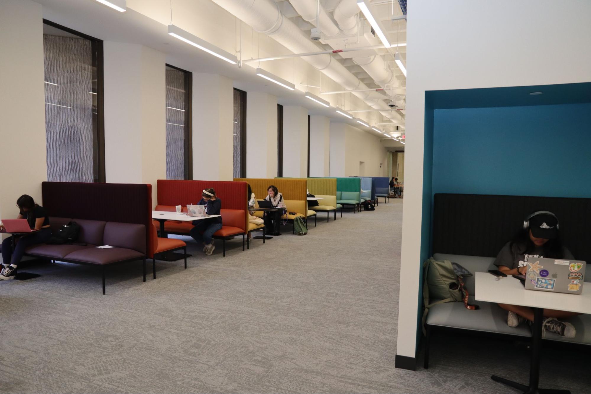 The open study space in the Scholars Lab features colorful couches near large windows. Additionally, there is a couch positioned on the right side of the colorful couches, adjacent to a wall. Individuals can be seen reading or using laptops.