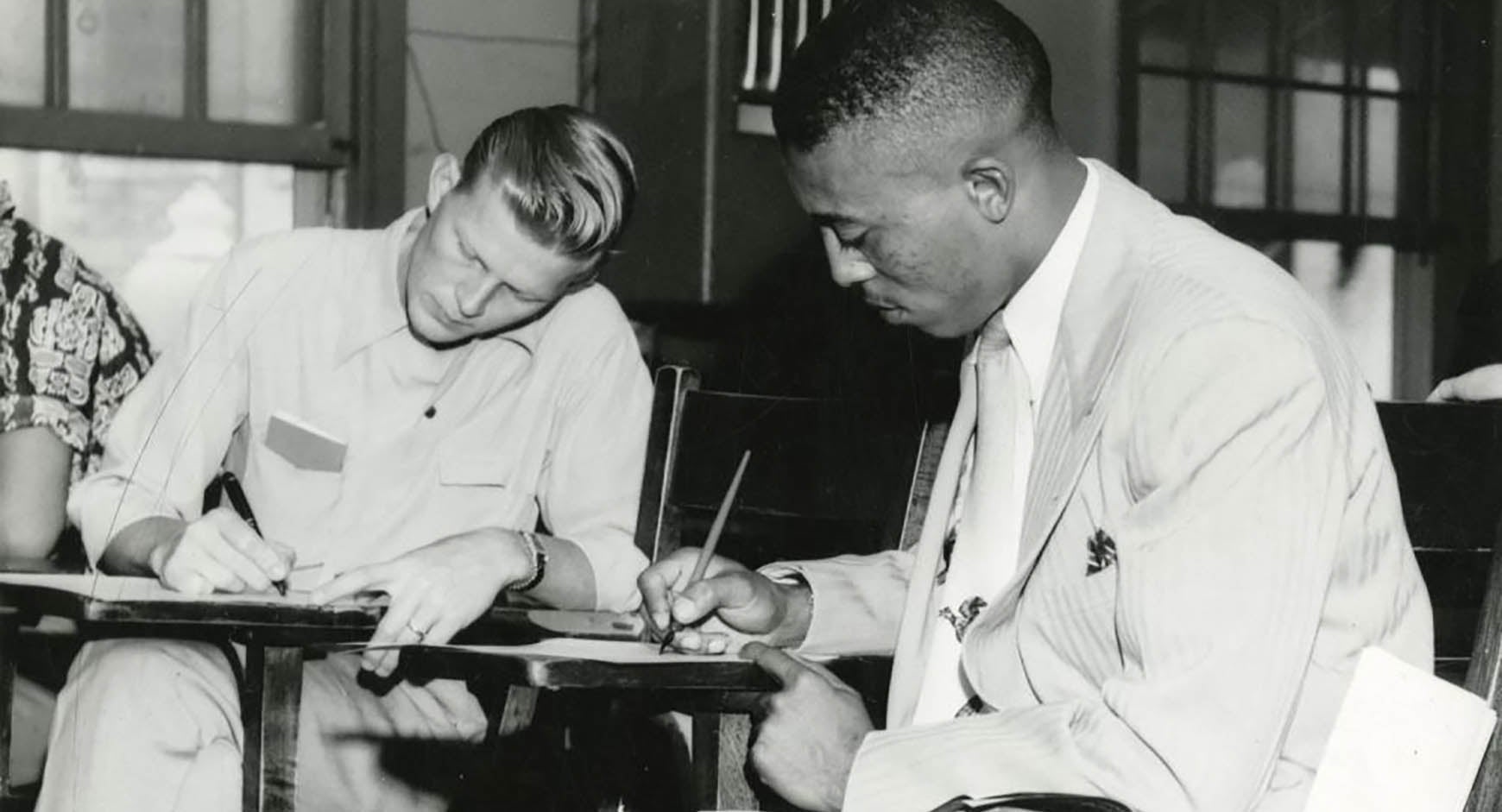 Black-and-white photo of John S. Chase, the first African American graduate of the University of Texas School of Architecture, seated at a desk and writing with a pen. He is wearing a light-colored suit and tie, focusing intently on his work. A fellow student is seated beside him, also writing, wearing a short-sleeved shirt. The setting appears to be a classroom, with desks and windows in the background.