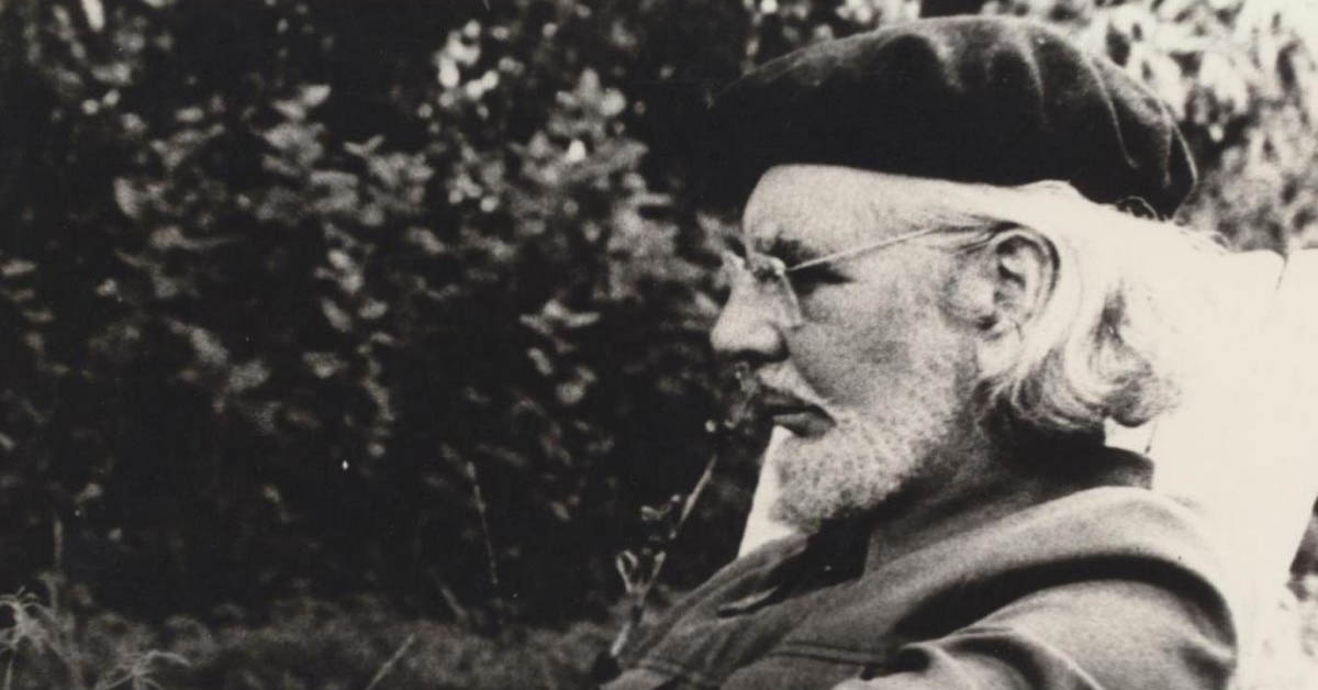 Black-and-white photograph of Ernesto Cardenal, a Nicaraguan poet and priest, sitting outdoors. He is wearing a beret and glasses, with his head turned to the side, showcasing his beard and long hair. The background features blurred foliage, creating a serene and reflective atmosphere.