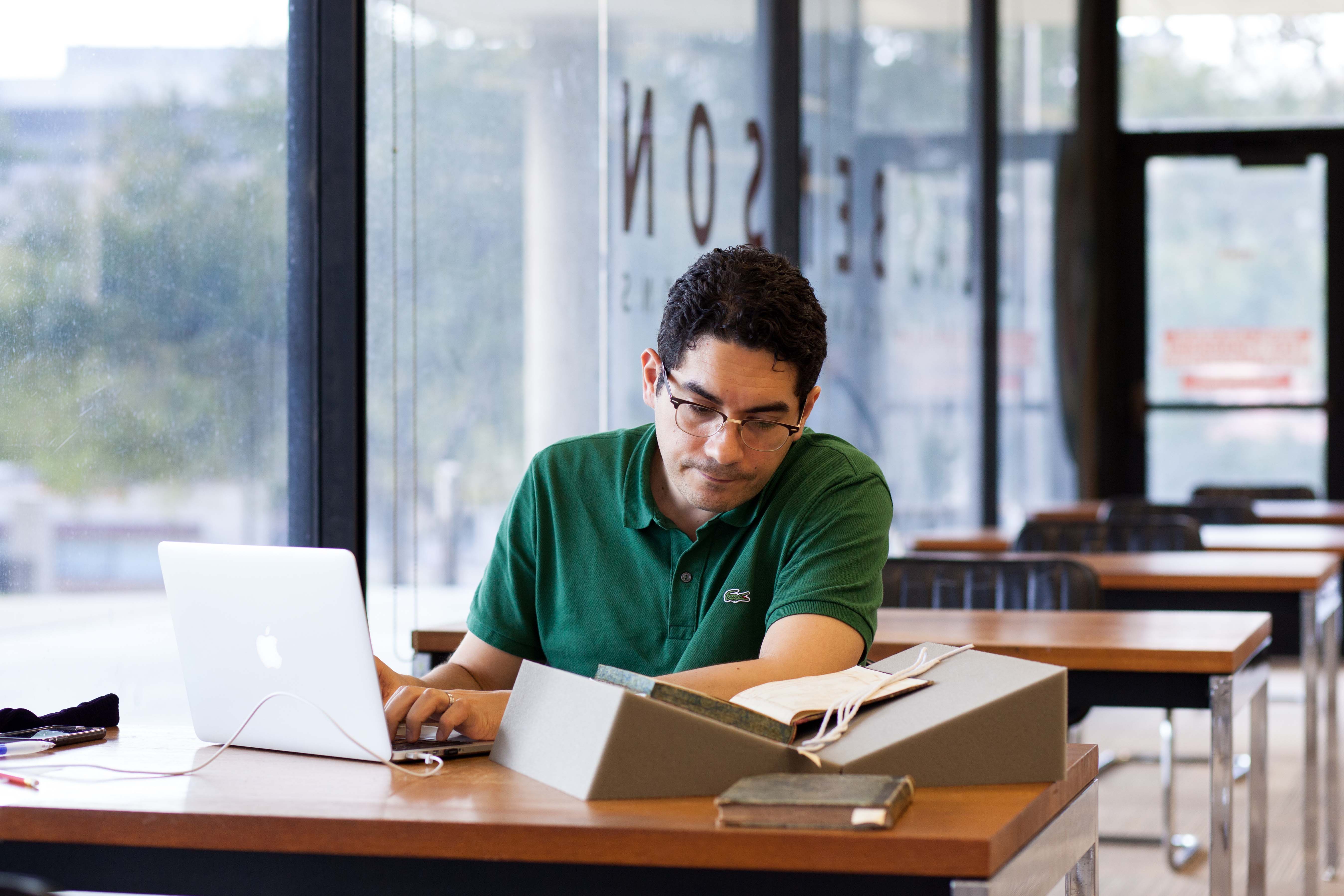 A man wearing a green polo shirt and glasses is seated at a wooden table in a library, working on a laptop while referencing an open archival book propped on a foam cradle. The setting is quiet, with natural light streaming through large windows and empty tables in the background.