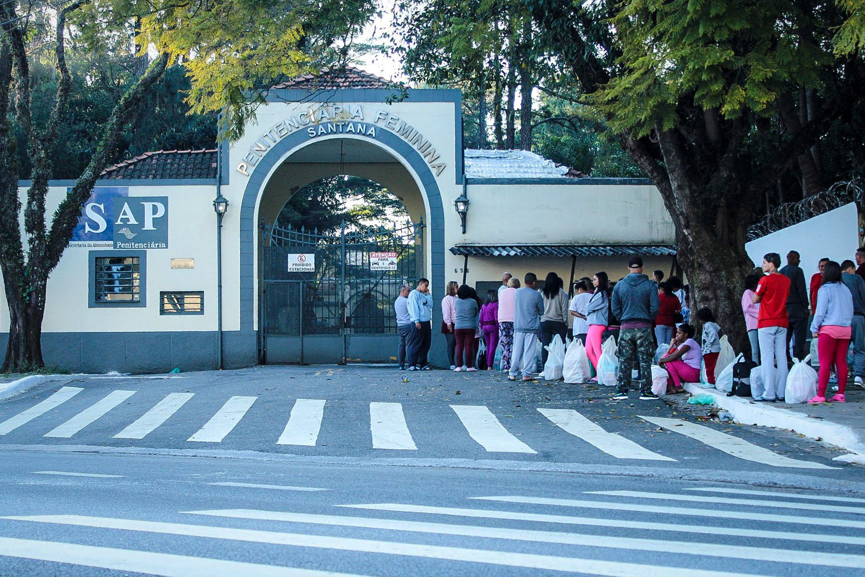 People stand in line outside the entrance gate of the Penitenciária Feminina de Santana, São Paulo’s largest women’s prison, holding bags likely filled with items for incarcerated loved ones. A sign for the São Paulo State Department of Penitentiary Administration (SAP) is visible on the prison wall. Trees frame the scene and a crosswalk leads to the gate.