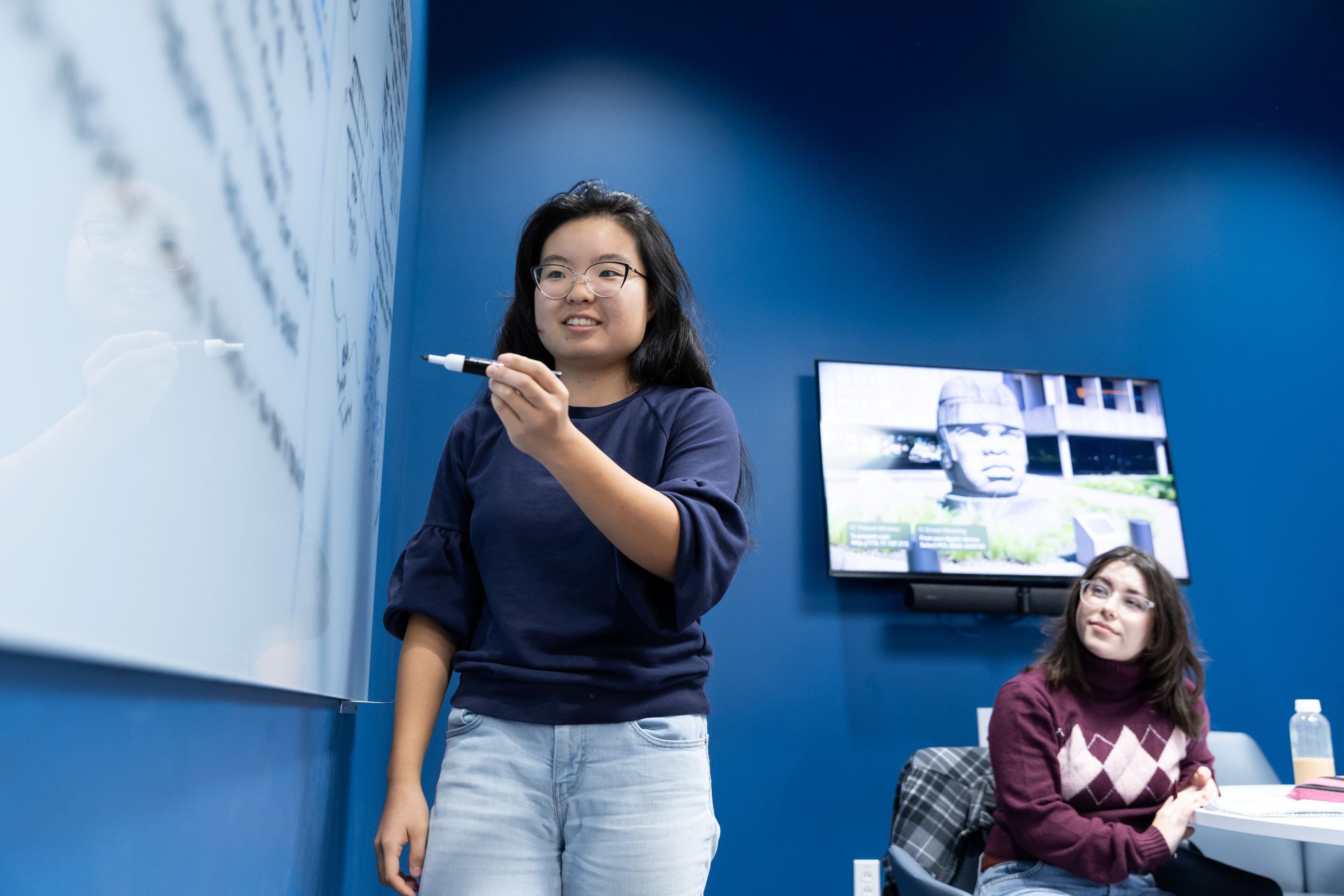 Two female students discuss at a white board.