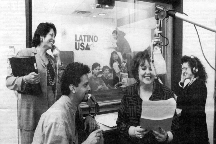 "A black-and-white photograph captures a lively moment inside a radio studio. In the foreground, a woman with short hair and a patterned blazer is smiling while reading from a script into a large studio microphone. To her left, a man with curly hair leans in, speaking into a microphone with a smile. Behind them, a woman wearing headphones listens attentively, adjusting them with one hand. Another woman, standing to the left, is holding a folder and laughing while speaking into a phone. Through the glass par