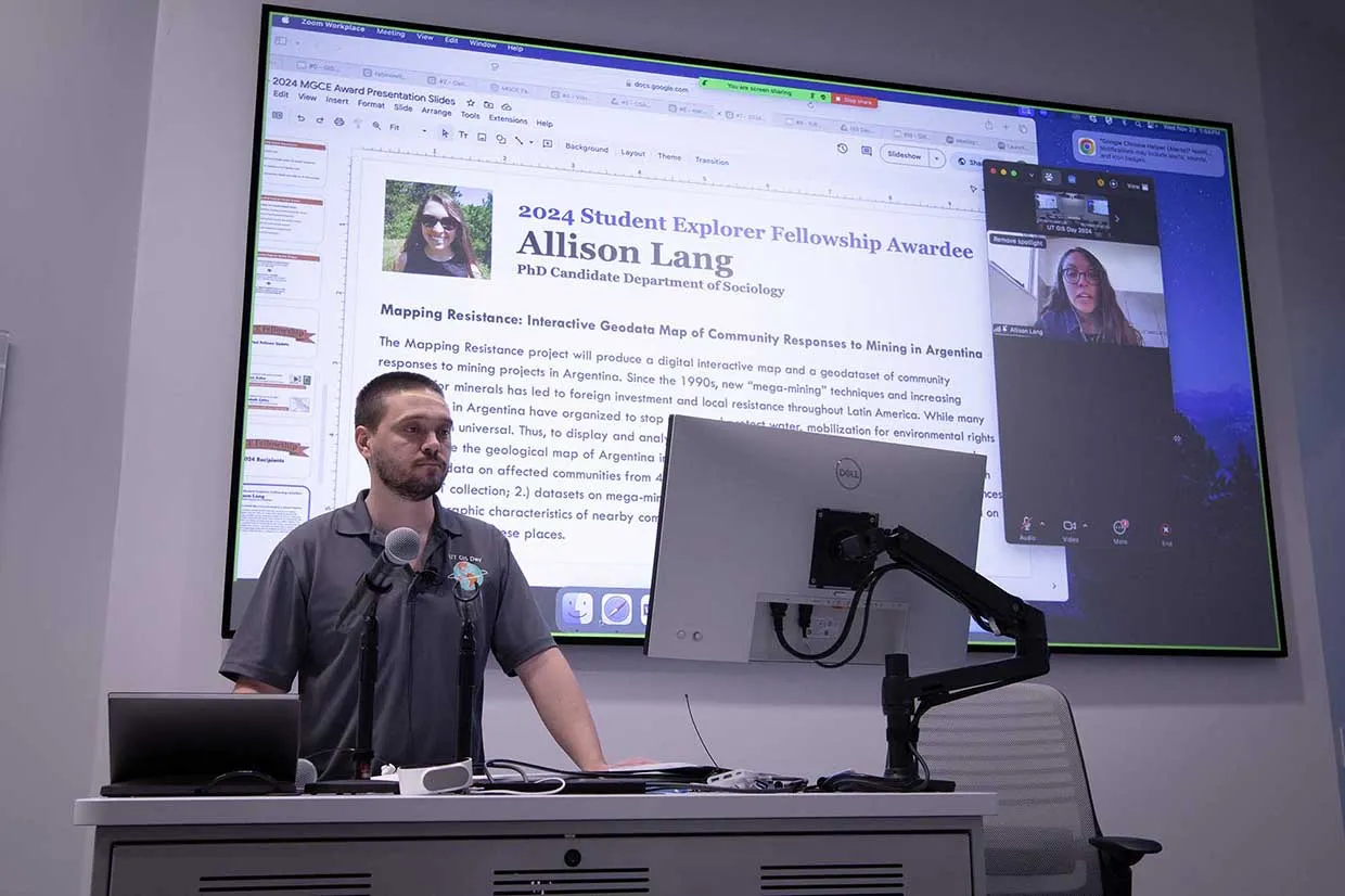 An individual stands at a podium equipped with microphones and a computer, presenting during an event. Behind him, a large screen displays information about the 2024 Student Explorer Fellowship awardee Allison Lang, a PhD candidate in the Department of Sociology, showcasing her project 'Mapping Resistance: Interactive Geodata Map of Community Responses to Mining in Argentina.' A video call featuring Allison Lang is visible on the screen.