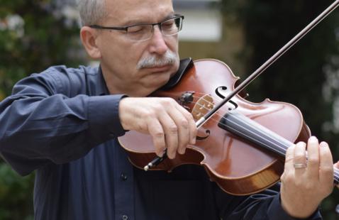 Dr. Domjan playing a viola, holding the instrument under the chin while drawing the bow across the strings, with greenery softly out of focus in the background
